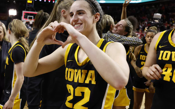 A HD wallpaper featuring a basketball player in a black and yellow Iowa jersey with the number 22 making a heart shape with her hands, smiling with her teammates in the background.