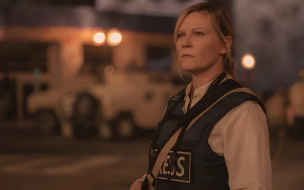 HD wallpaper featuring Kirsten Dunst in character, with a serious expression, wearing a vest with PRESS across the front against a dusk-lit backdrop with military vehicles, suggesting a scene from the movie Civil War (2024).
