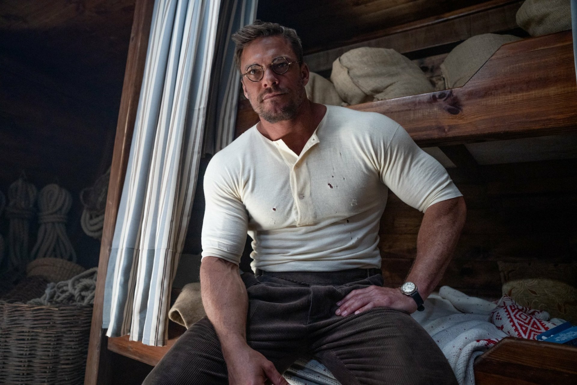 HD wallpaper featuring a character from the movie The Ministry of Ungentlemanly Warfare sitting thoughtfully, portrayed by an actor wearing a fitted white shirt and brown trousers.