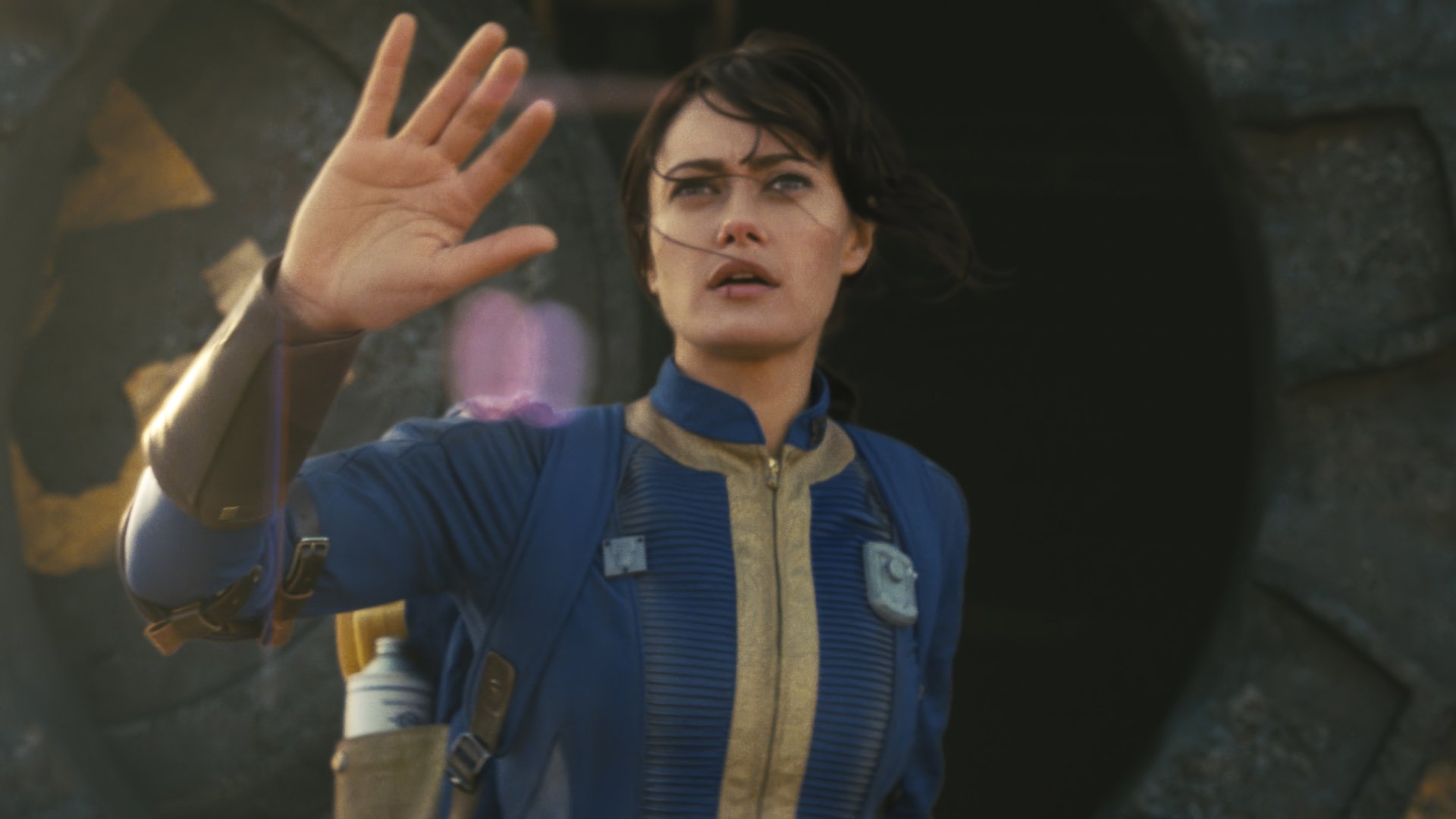 HD desktop wallpaper from the TV show Fallout featuring Ella Purnell in a blue jumpsuit, extending her hand with a futuristic glow.