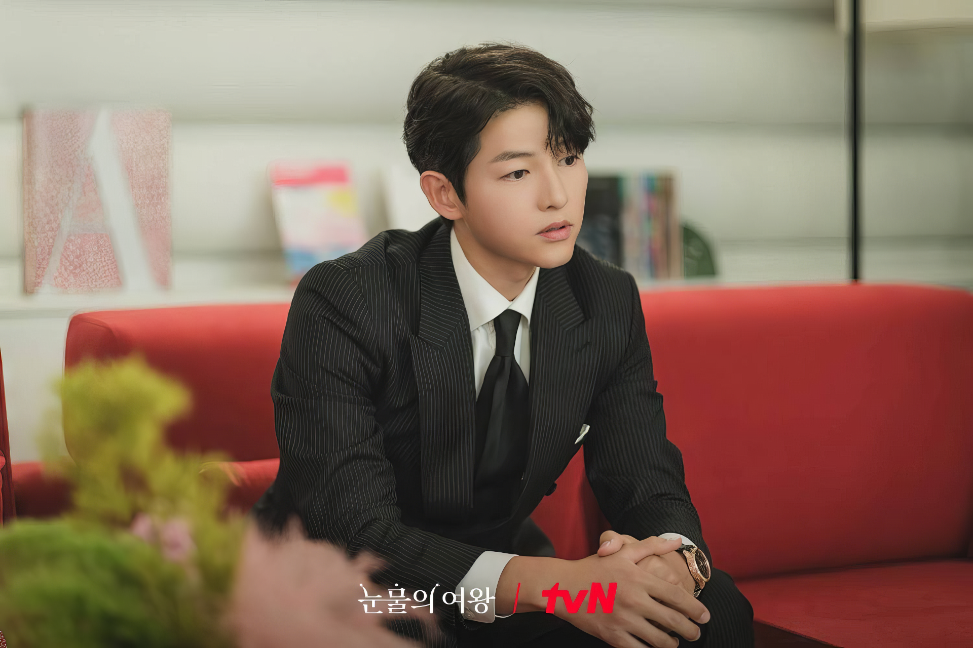 HD wallpaper of a stylish young man in a pinstripe suit sitting on a red sofa, from the TV show Queen of Tears on tvN.