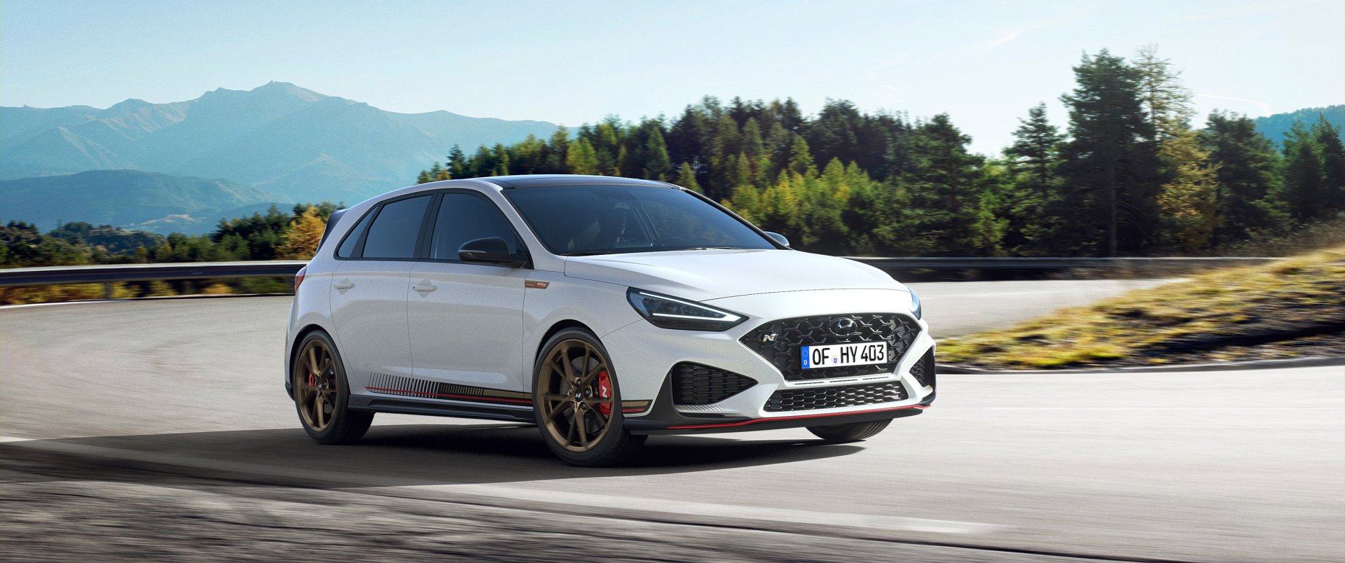 A high-definition desktop wallpaper featuring a white Hyundai i30 driving on a road with scenic forest and mountain backdrop. The vehicle showcases sleek lines and sporty design.