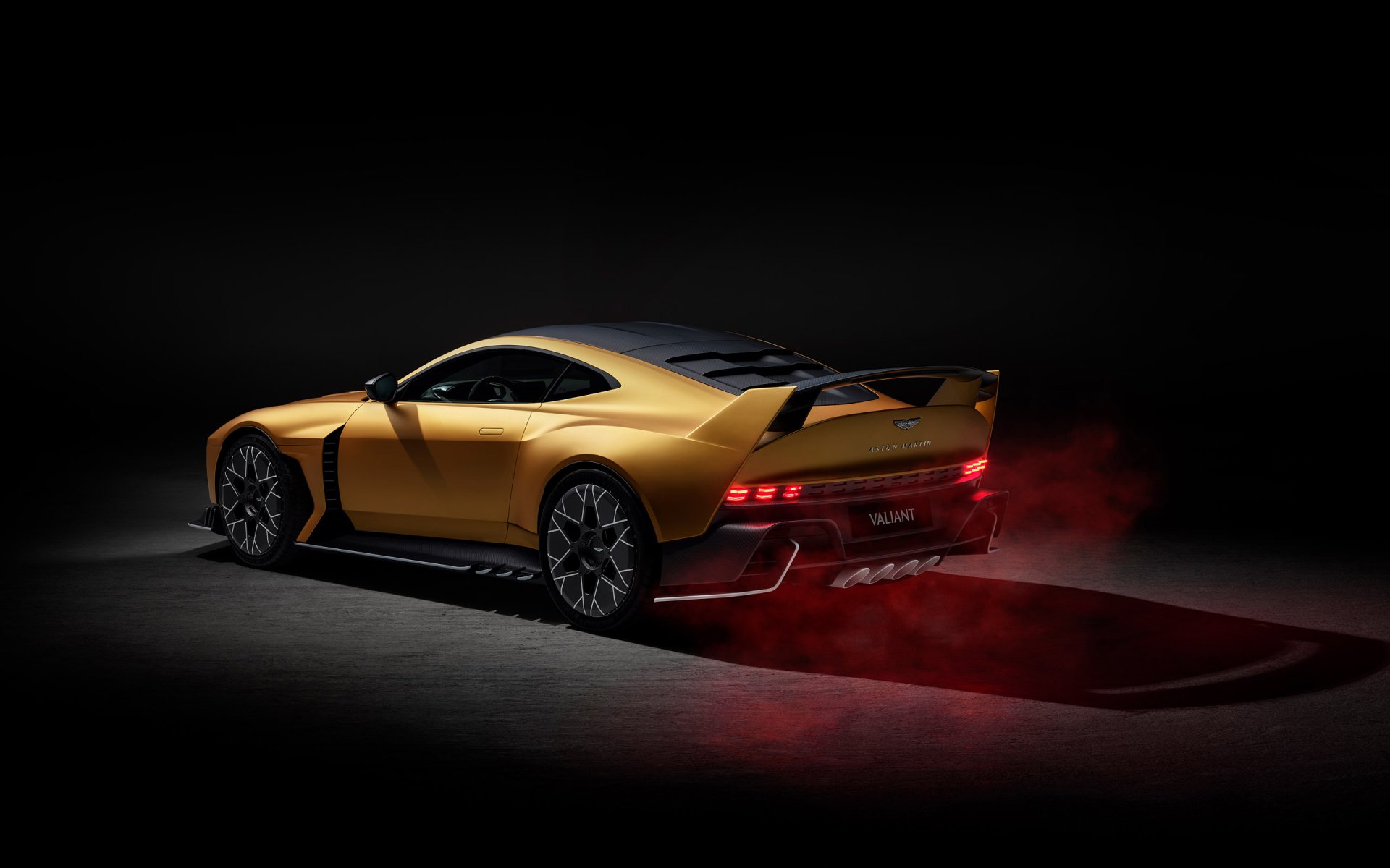 HD desktop wallpaper featuring an Aston Martin Valiant, a sleek yellow sports car with a sporty design, captured in a low light setting with red brake lights illuminating the dark background.