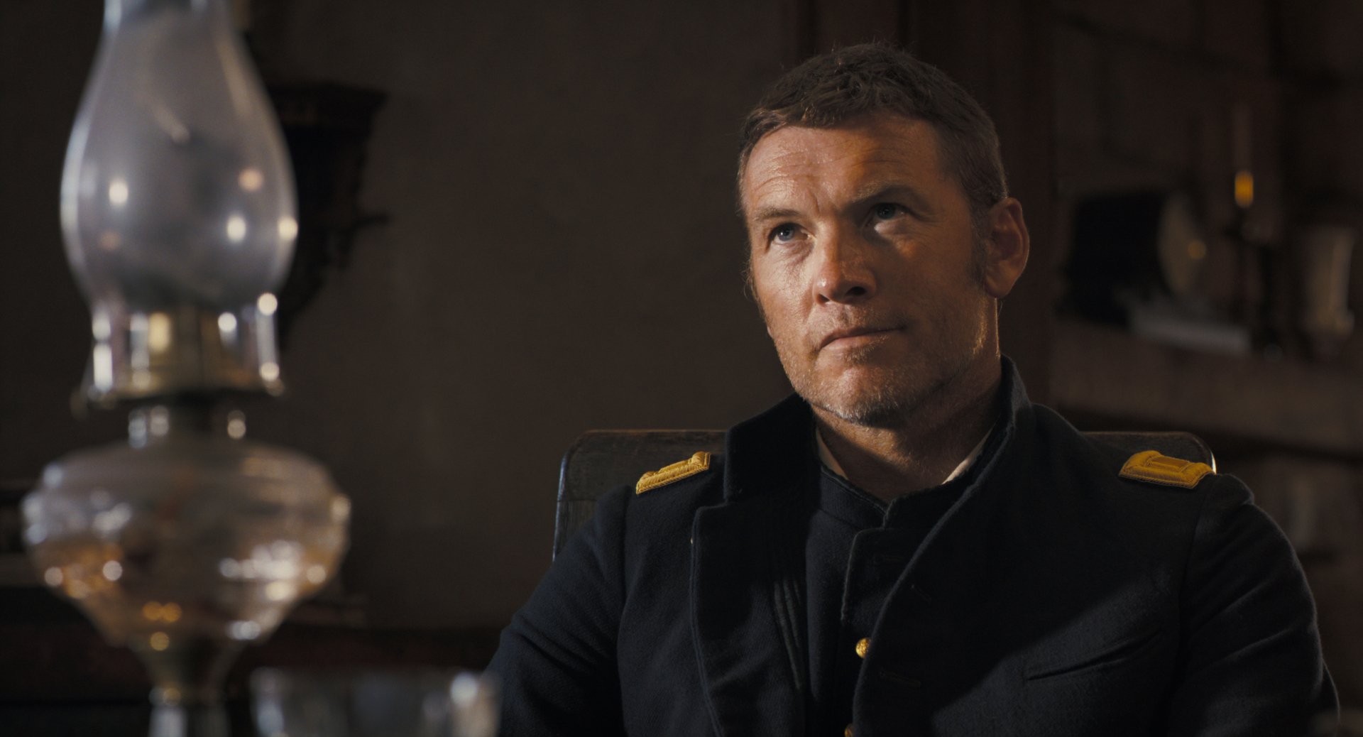 HD wallpaper featuring Sam Worthington in character from the movie Horizon: An American Saga - Chapter 1, seated and wearing a dark uniform with yellow insignia.