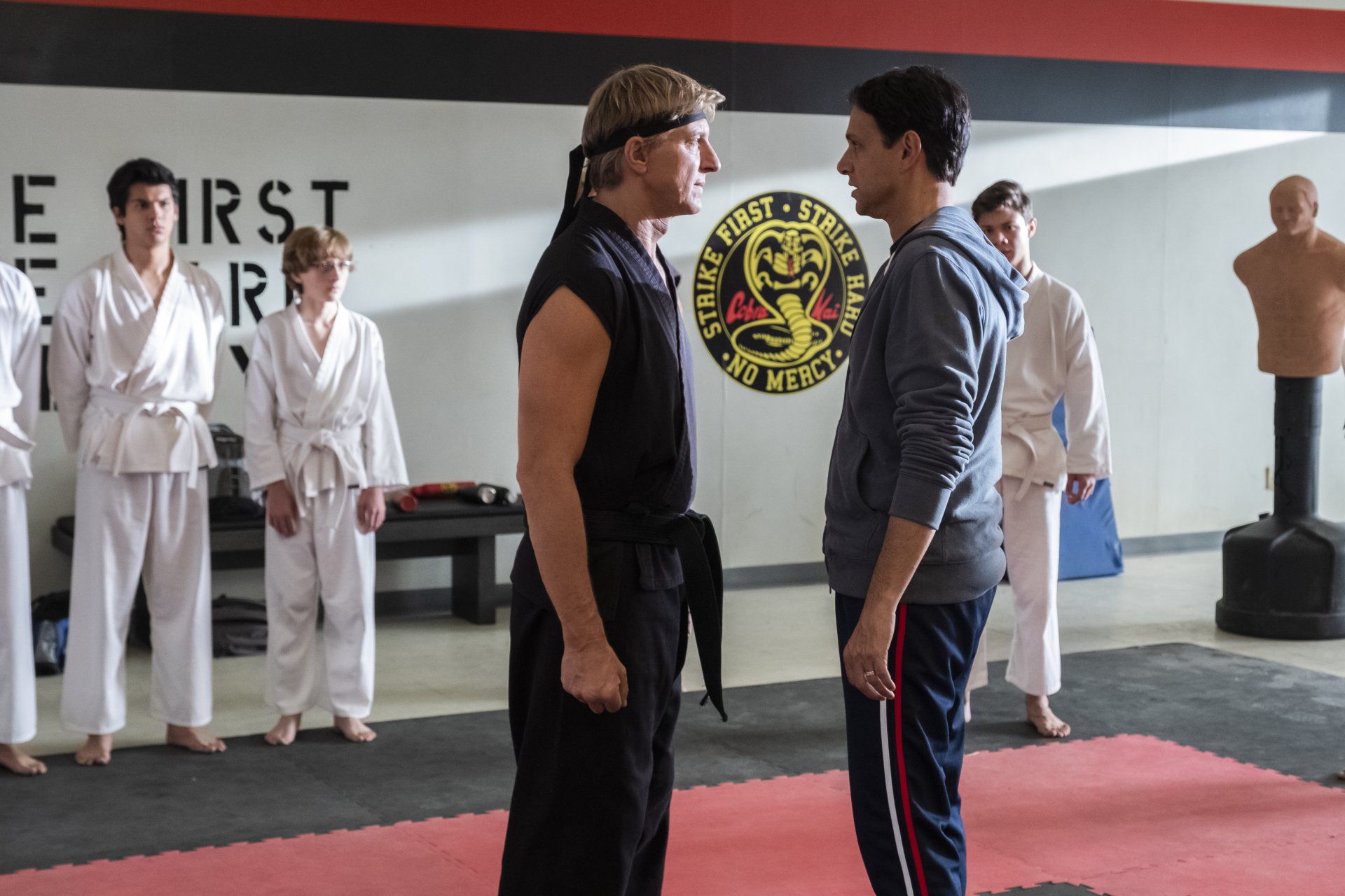 HD desktop wallpaper featuring a tense face-off between two characters from the TV show Cobra Kai, with students in white gis observing in the background.