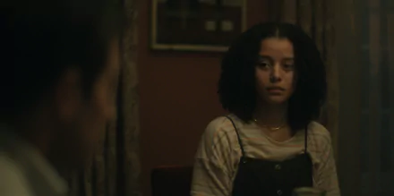 A HD image from the TV show Presumed Innocent (2024), featuring a girl with curly hair sitting in a dimly lit room, in a contemplative moment with another character partially visible.