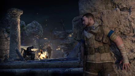 A high-definition desktop wallpaper and background from the video game Sniper Elite III, showing a soldier in tactical gear hiding behind ancient ruins, observing enemies by a campfire at night.