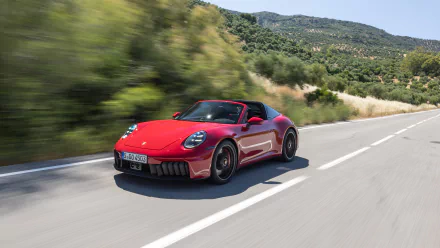 HD wallpaper of a red Porsche 911 Targa 4 GTS cruising on a scenic road, surrounded by greenery and hills in the background.