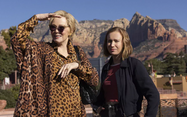 Two women stand outdoors against a backdrop of mountains, showcasing a scene from the TV show Hacks (2021). One woman wears a leopard-print coat, while the other sports a casual look.