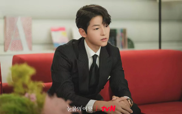 HD wallpaper of a stylish young man in a pinstripe suit sitting on a red sofa, from the TV show Queen of Tears on tvN.
