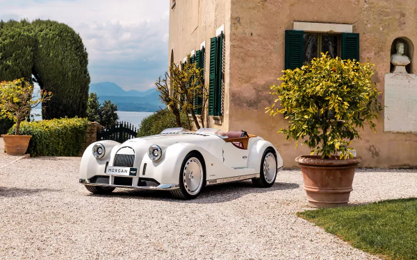 HD desktop wallpaper featuring a Morgan Midsummer parked in a picturesque courtyard with lush greenery and a scenic lake in the background.