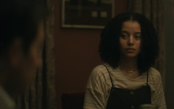 A HD image from the TV show Presumed Innocent (2024), featuring a girl with curly hair sitting in a dimly lit room, in a contemplative moment with another character partially visible.