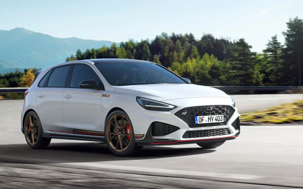 A high-definition desktop wallpaper featuring a white Hyundai i30 driving on a road with scenic forest and mountain backdrop. The vehicle showcases sleek lines and sporty design.