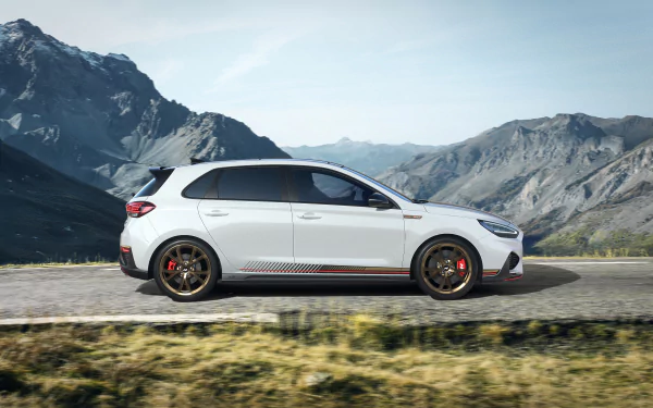 HD desktop wallpaper featuring a white Hyundai i30 parked on a mountain road, with a stunning backdrop of rocky hills and clear blue skies.