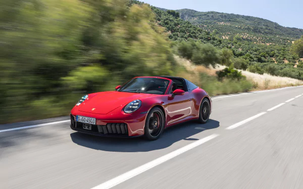 HD wallpaper of a red Porsche 911 Targa 4 GTS cruising on a scenic road, surrounded by greenery and hills in the background.
