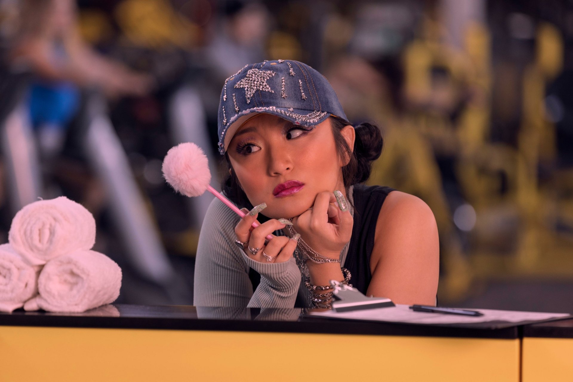 A still from the movie Space Cadet, featuring a contemplative character wearing a denim cap, resting her chin on her hand, set against a vibrant gym backdrop.