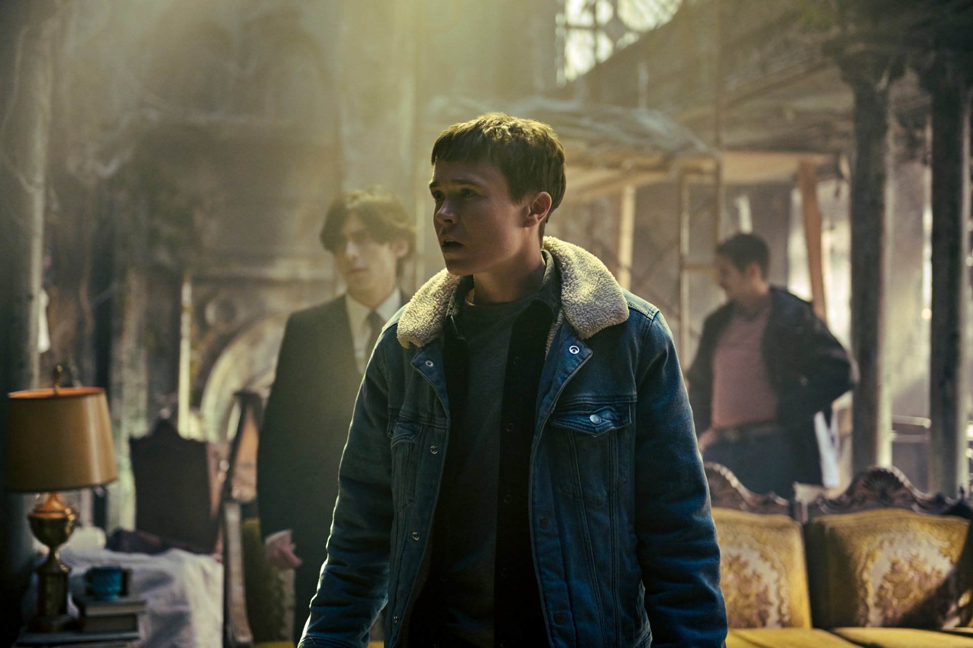 A high-definition wallpaper from The Umbrella Academy, featuring a tense scene with a young man in a denim jacket and two others in a dimly lit, seemingly abandoned room.