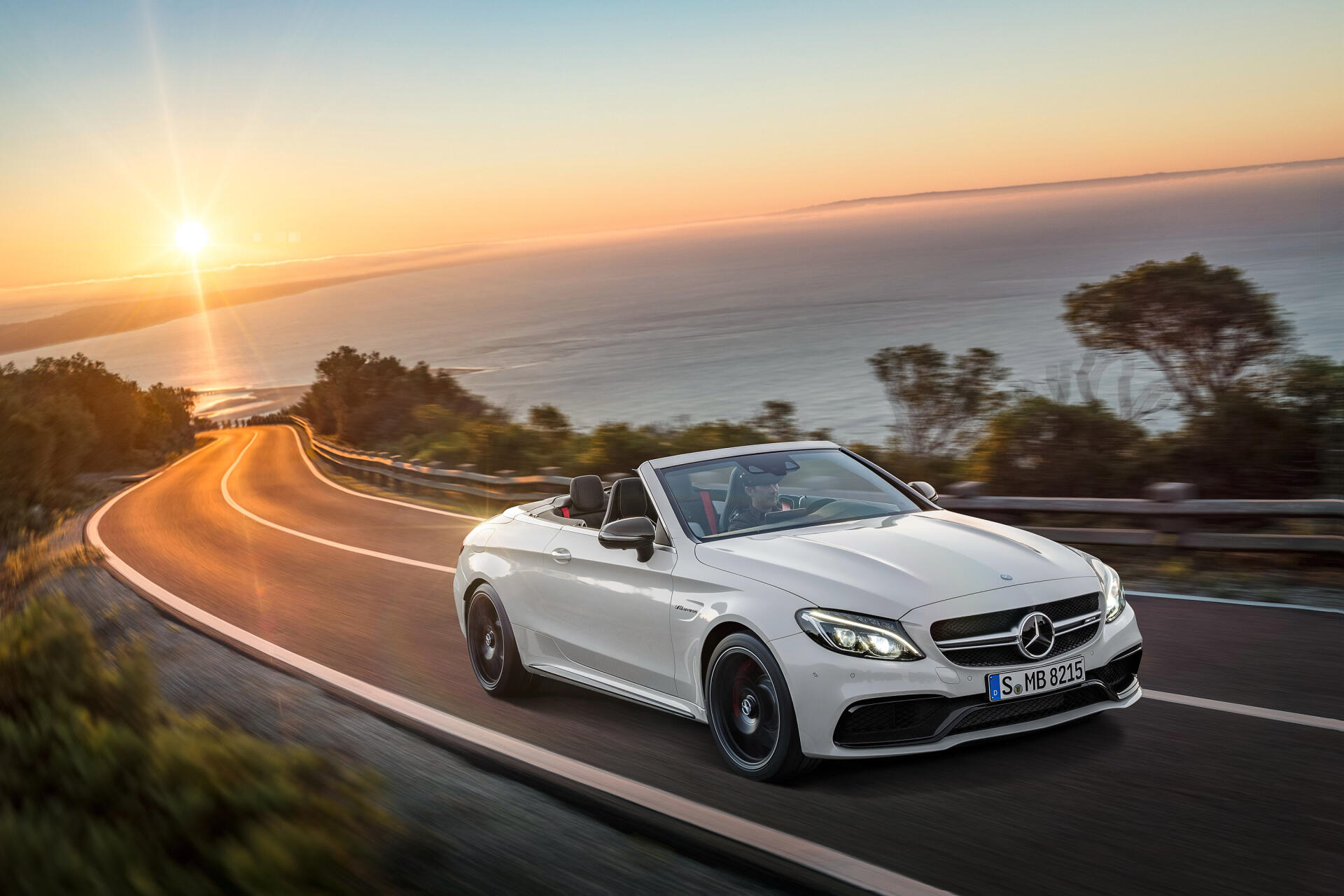 A sleek white Mercedes-Benz C63 AMG convertible cruises along a coastal road at sunset, showcasing its luxury and performance in this stunning HD desktop wallpaper.