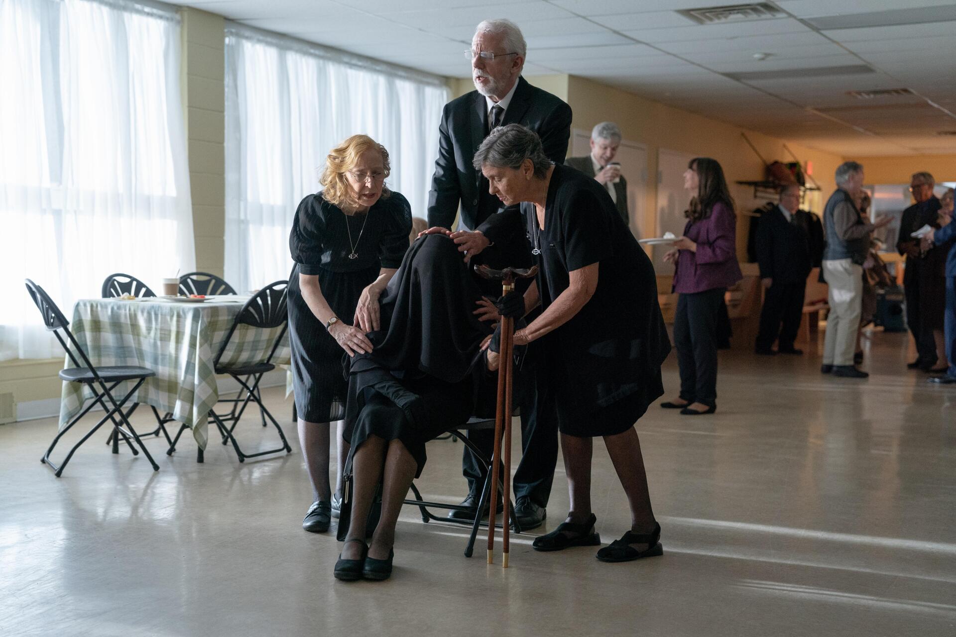 A scene from the movie The Front Room, featuring characters engaged in a heartfelt moment, set in a bright, spacious room with folding chairs and other attendees in the background.