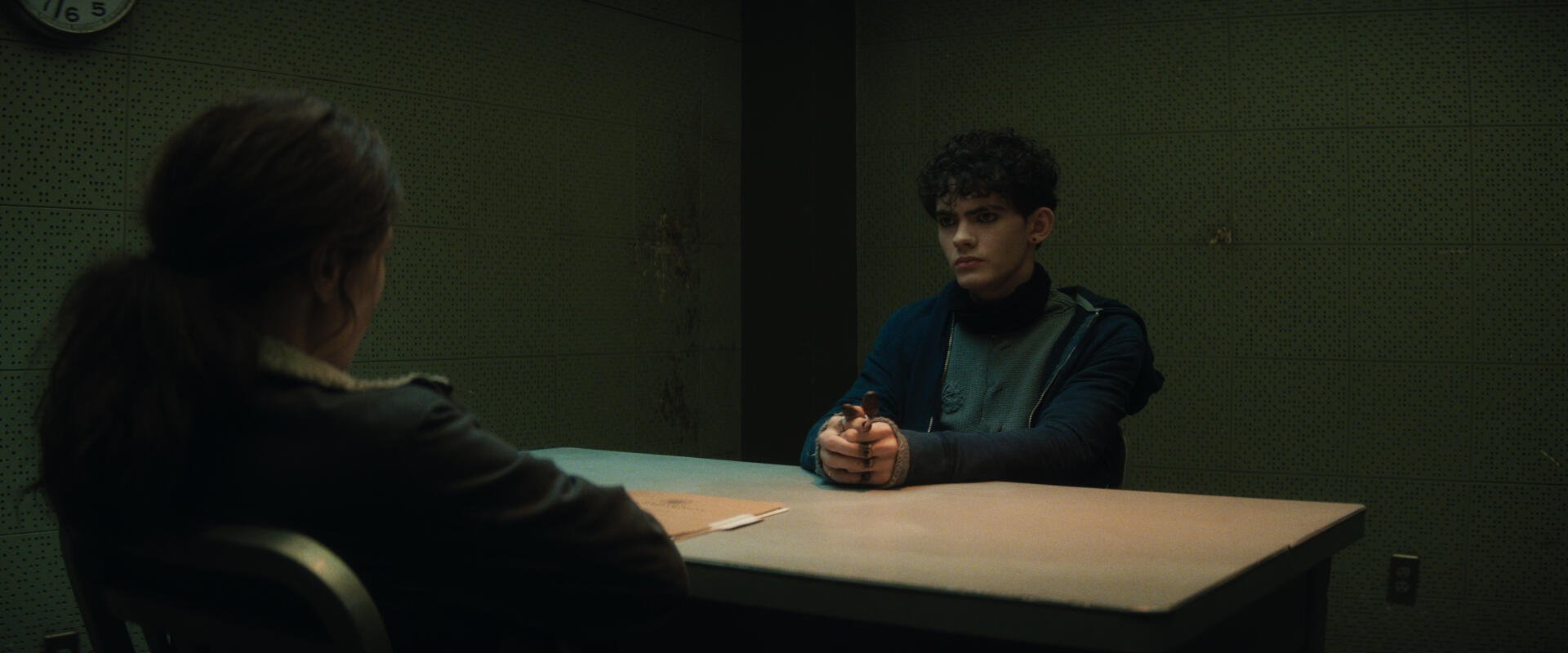 A tense scene from the TV show Agatha All Along, featuring two characters in a dimly lit interrogation room, with one seated at a table, holding a weapon. HD wallpaper background.