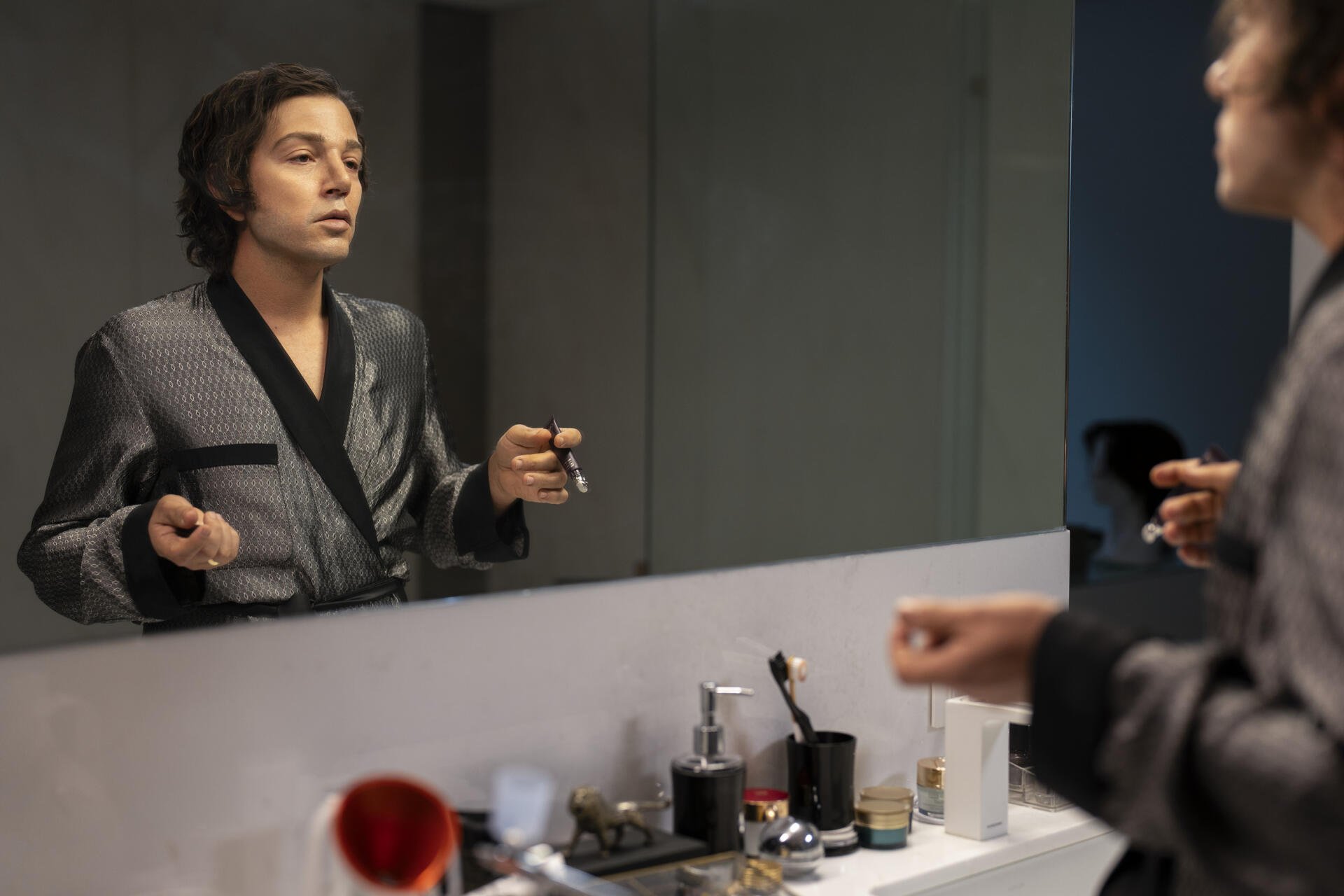 A scene from the TV show La Máquina (2024), featuring a character in a robe applying makeup while gazing into a mirror, surrounded by various beauty products.