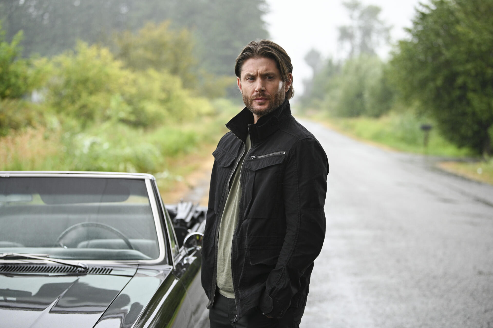 A high-definition desktop wallpaper featuring Jensen Ackles from the TV show Tracker (2024), standing beside a vintage car on a serene, tree-lined road.