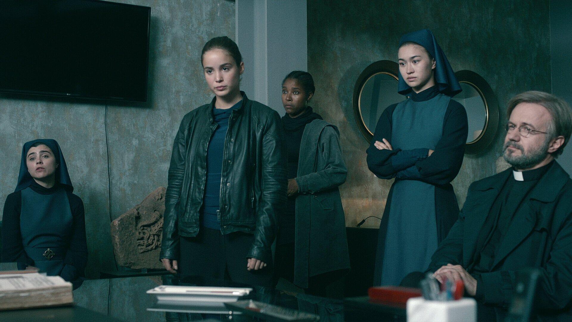 A scene from the TV show Warrior Nun, featuring Alba Baptista and fellow cast members in a tense gathering, set against a modern, minimalistic background.