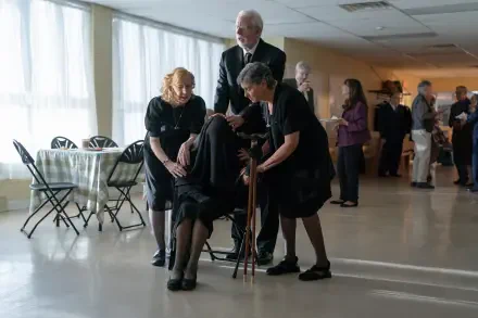 A scene from the movie The Front Room, featuring characters engaged in a heartfelt moment, set in a bright, spacious room with folding chairs and other attendees in the background.