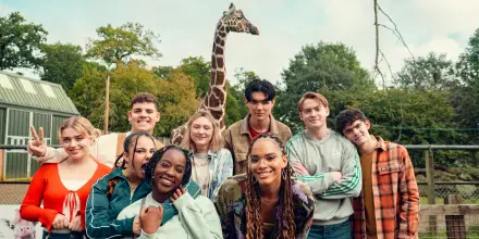 A lively group photo from the TV show *Heartstopper*, featuring characters smiling together with a giraffe in the background, set against a green, natural landscape.