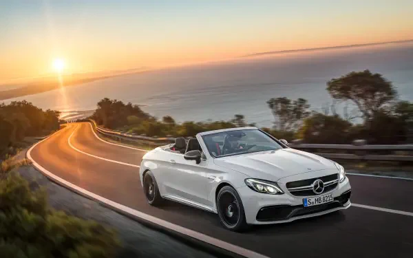 A sleek white Mercedes-Benz C63 AMG convertible cruises along a coastal road at sunset, showcasing its luxury and performance in this stunning HD desktop wallpaper.