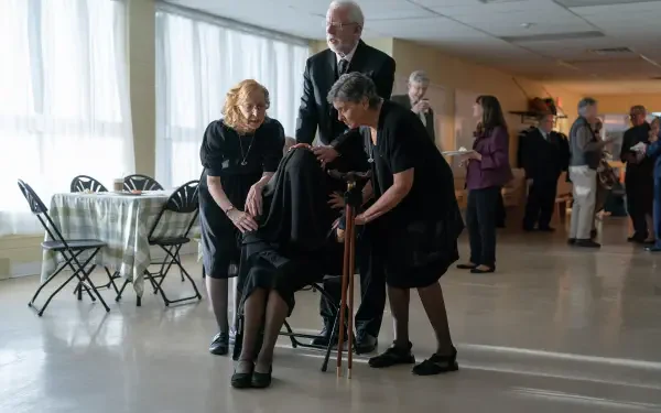 A scene from the movie The Front Room, featuring characters engaged in a heartfelt moment, set in a bright, spacious room with folding chairs and other attendees in the background.