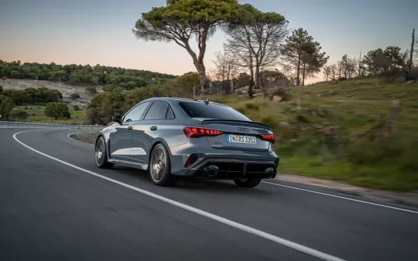 A sleek Audi RS3 Sedan drives along a winding road, surrounded by greenery, showcasing its dynamic design in this vibrant 4K Ultra HD desktop wallpaper.