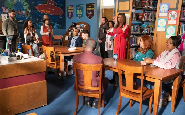 A vibrant scene from the TV show *Abbott Elementary*, featuring a diverse group of characters gathered in a classroom, engaged in discussion around wooden tables, with bookshelves in the background.