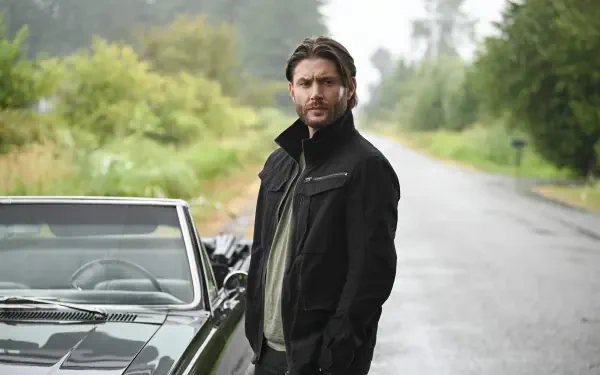 A high-definition desktop wallpaper featuring Jensen Ackles from the TV show Tracker (2024), standing beside a vintage car on a serene, tree-lined road.
