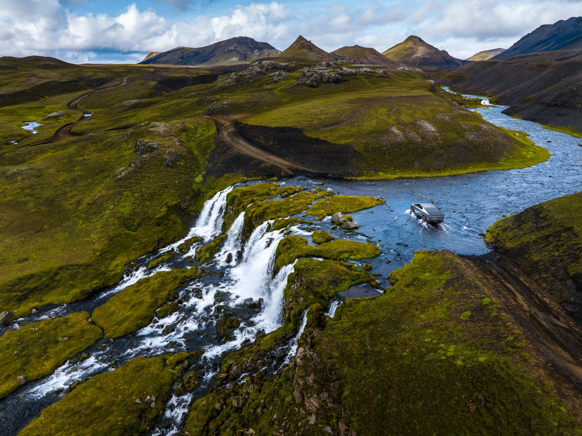 A stunning 4K Ultra HD wallpaper featuring a Tesla Cybertruck navigating through a breathtaking landscape with waterfalls and flowing rivers, surrounded by lush greenery and mountains.