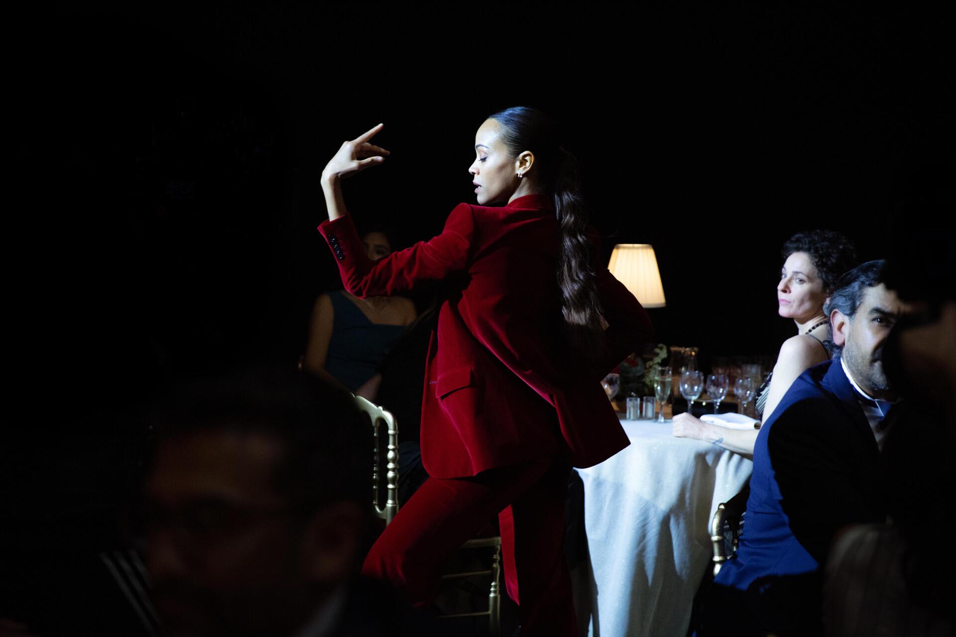 4K Ultra HD movie wallpaper featuring Emilia Pérez in a striking red outfit performing, with Zoe Saldana and others seated at a dimly lit table in the background.