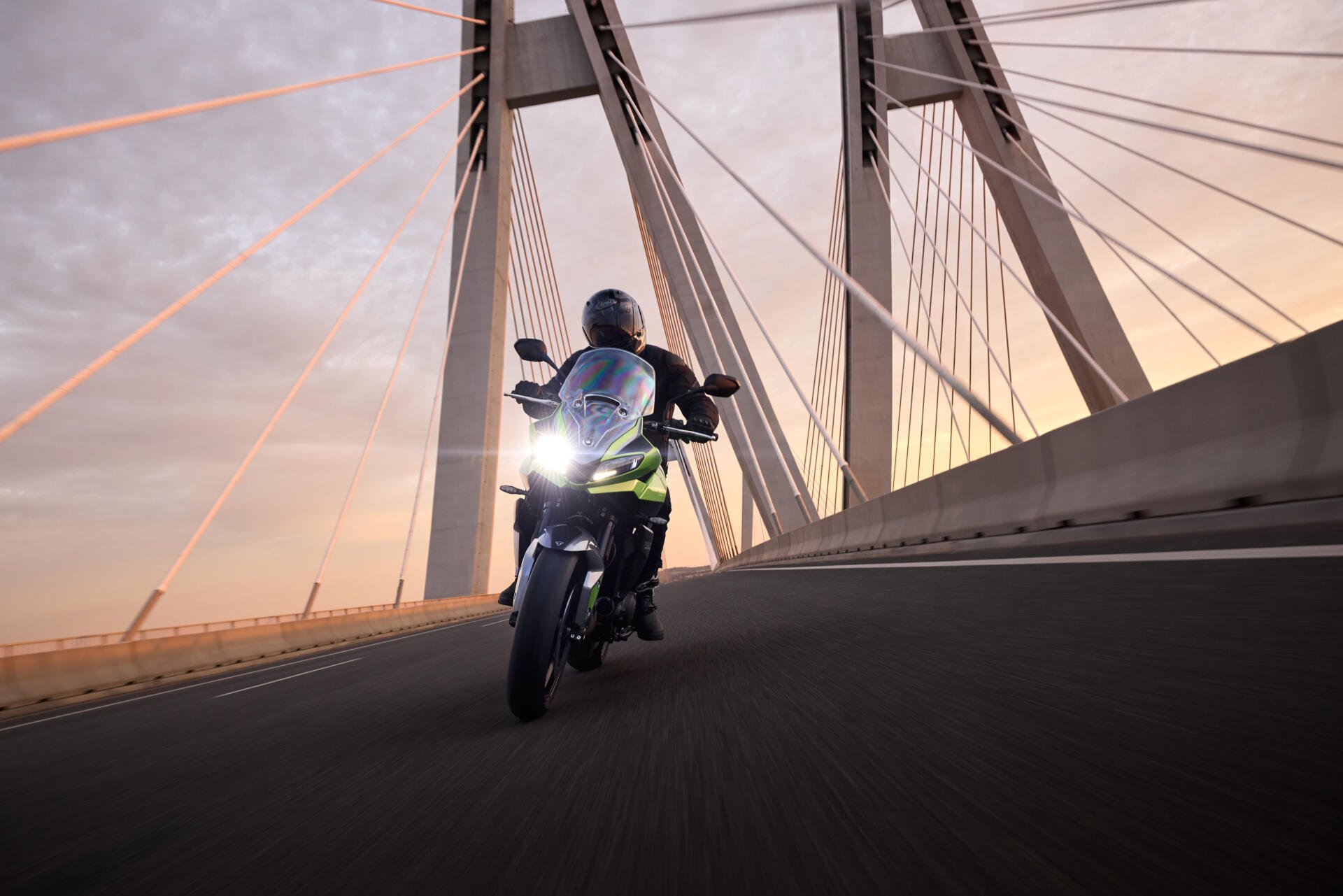 A Triumph Tiger Sport 660 motorcycle glides along a bridge at sunset, showcasing sleek design and dynamic motion against an urban backdrop.