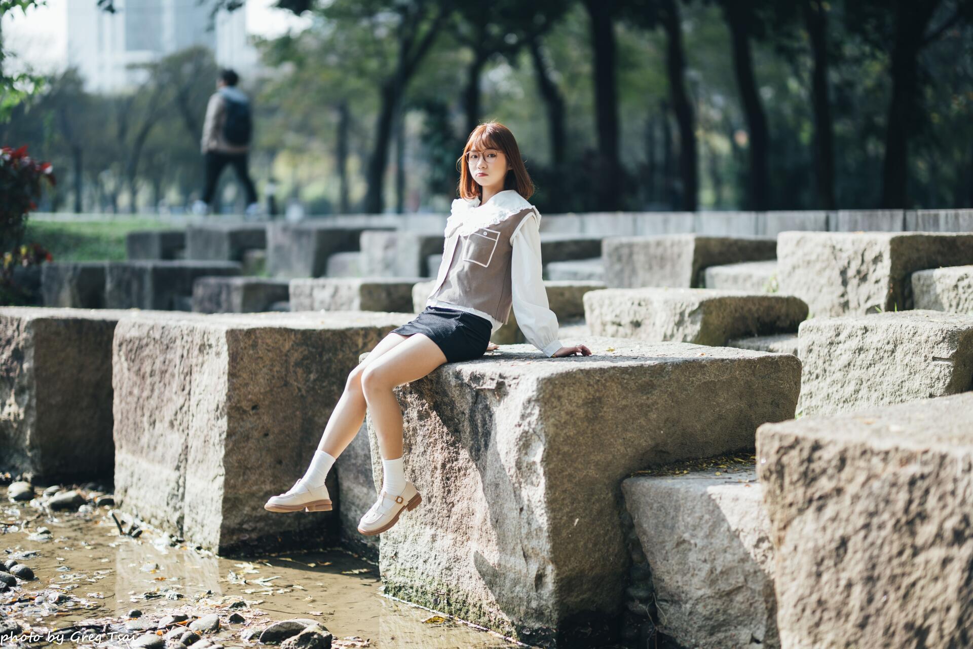 8K Ultra HD PC desktop wallpaper of an Asian woman sitting on stone blocks in a sunlit park, wearing a white blouse and skirt, serene urban greenery in soft bokeh background.