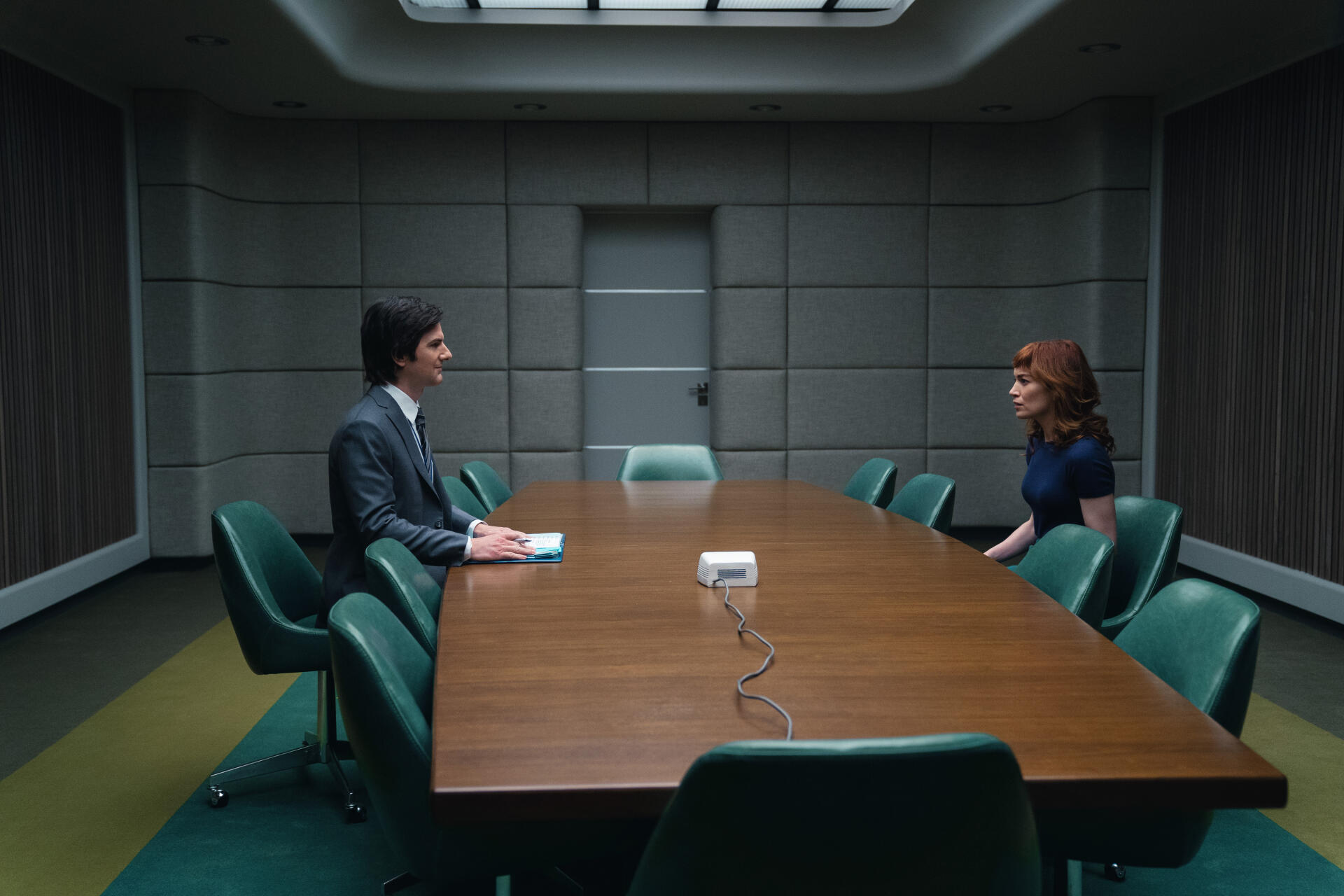 A scene from the TV show Severance featuring Britt Lower and Adam Scott sitting at a long conference table in a minimalist, modern room. 4K Ultra HD wallpaper.