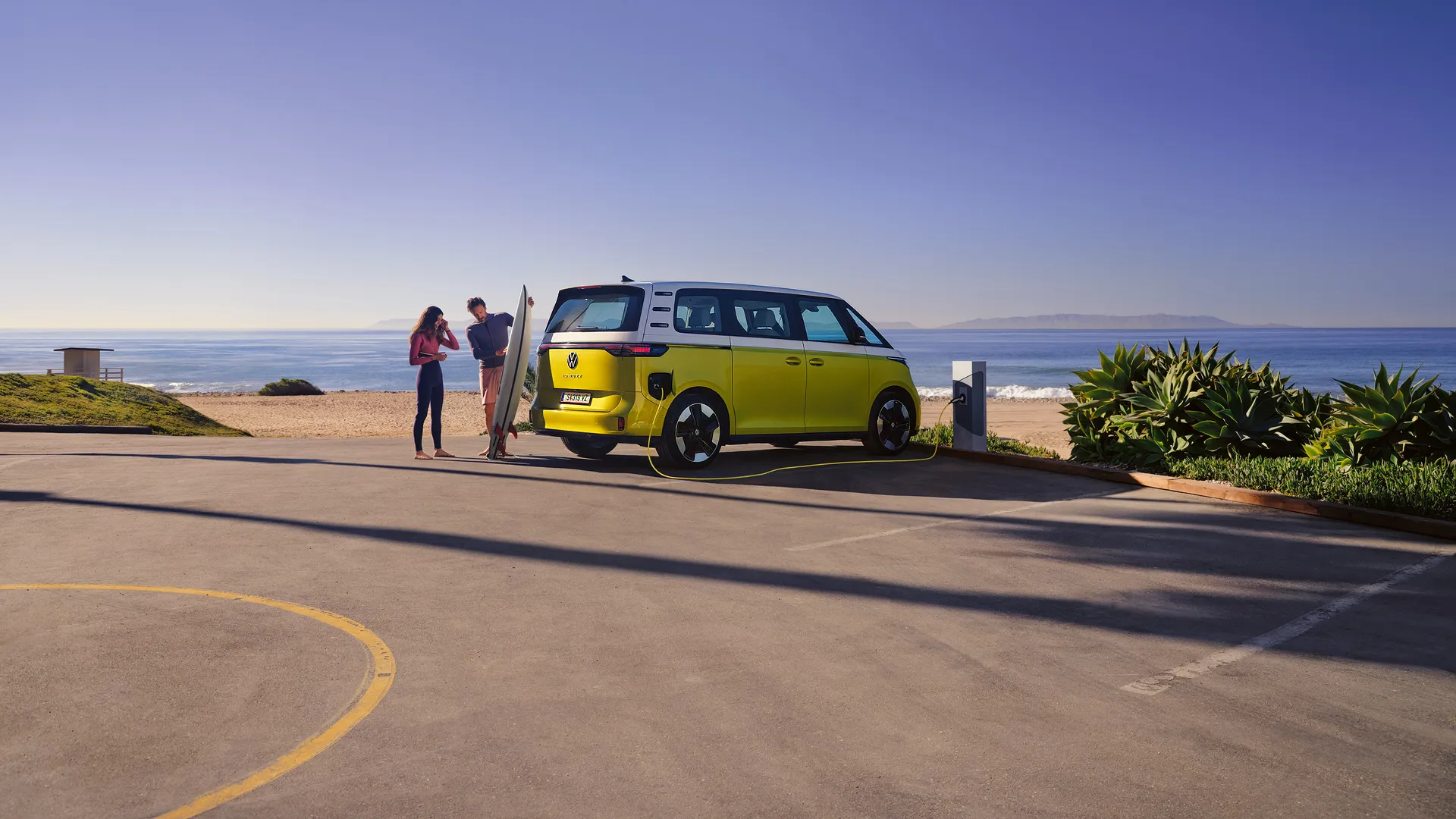 A vibrant yellow Volkswagen ID. Buzz electric car parked by the beach, with two people preparing to surf. The scene captures a sunny coastal atmosphere.