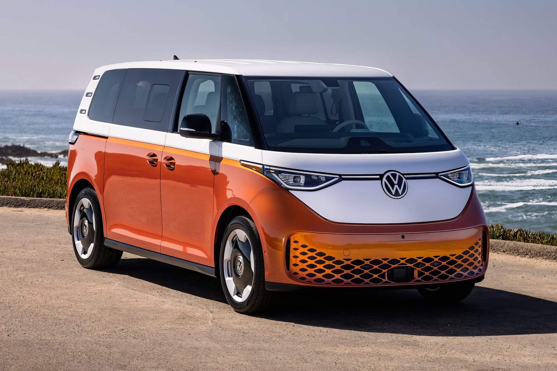 An orange and white Volkswagen ID. Buzz electric car parked by the coast, showcasing its modern design against a scenic ocean background.