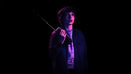 Finn Wolfhard as Mike Wheeler from Stranger Things stands in a dark setting, holding a radio, illuminated by a soft purple light. This HD image captures a suspenseful moment from the show.