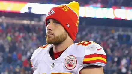 Travis Kelce of the Kansas City Chiefs stands on the field, wearing a vibrant Chiefs beanie, surrounded by a lively NFL atmosphere. A striking HD desktop wallpaper for football fans.