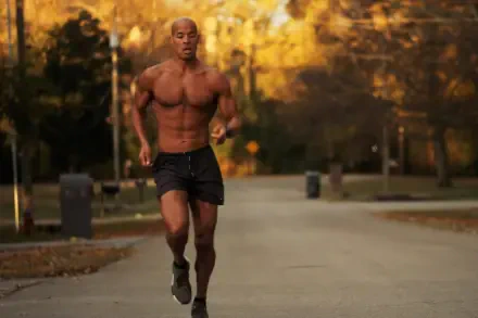 A muscular David Goggins runs through a park during autumn, showcasing determination and athleticism. This HD image serves as an inspiring desktop wallpaper for sports enthusiasts.