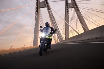 A Triumph Tiger Sport 660 motorcycle glides along a bridge at sunset, showcasing sleek design and dynamic motion against an urban backdrop.
