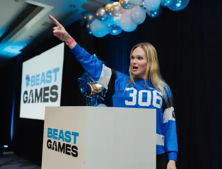 A lively host in a blue jersey excitedly gestures while presenting at the Beast Games event, with balloons and a backdrop showcasing the show's branding, set in a vibrant venue.