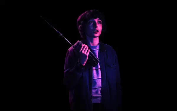 Finn Wolfhard as Mike Wheeler from Stranger Things stands in a dark setting, holding a radio, illuminated by a soft purple light. This HD image captures a suspenseful moment from the show.