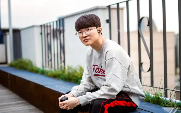 A high-definition wallpaper featuring T1's Faker, a prominent League of Legends player, casually sitting outdoors, sporting a Fake sweatshirt and looking at the camera.