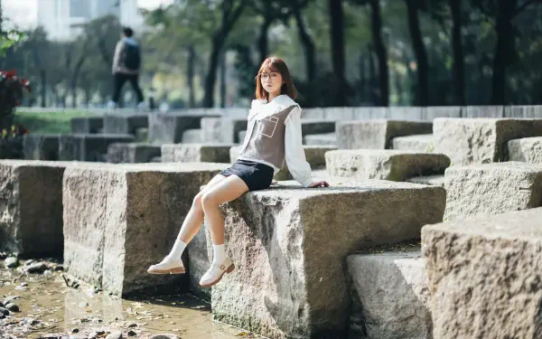 8K Ultra HD PC desktop wallpaper of an Asian woman sitting on stone blocks in a sunlit park, wearing a white blouse and skirt, serene urban greenery in soft bokeh background.