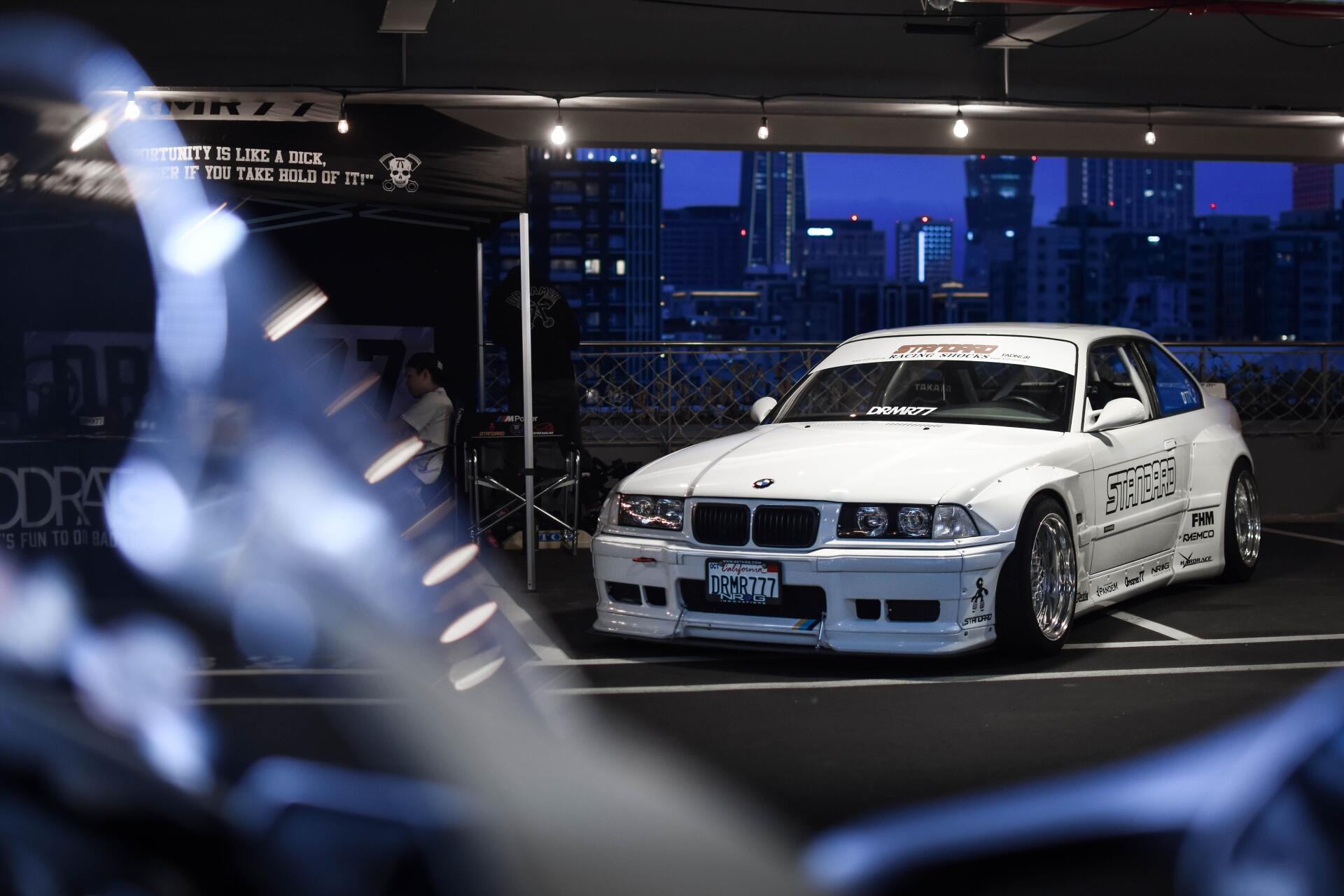 White BMW E36 race-styled car parked in a dim garage with city skyline at night — 5K Ultra HD PC desktop wallpaper/background.